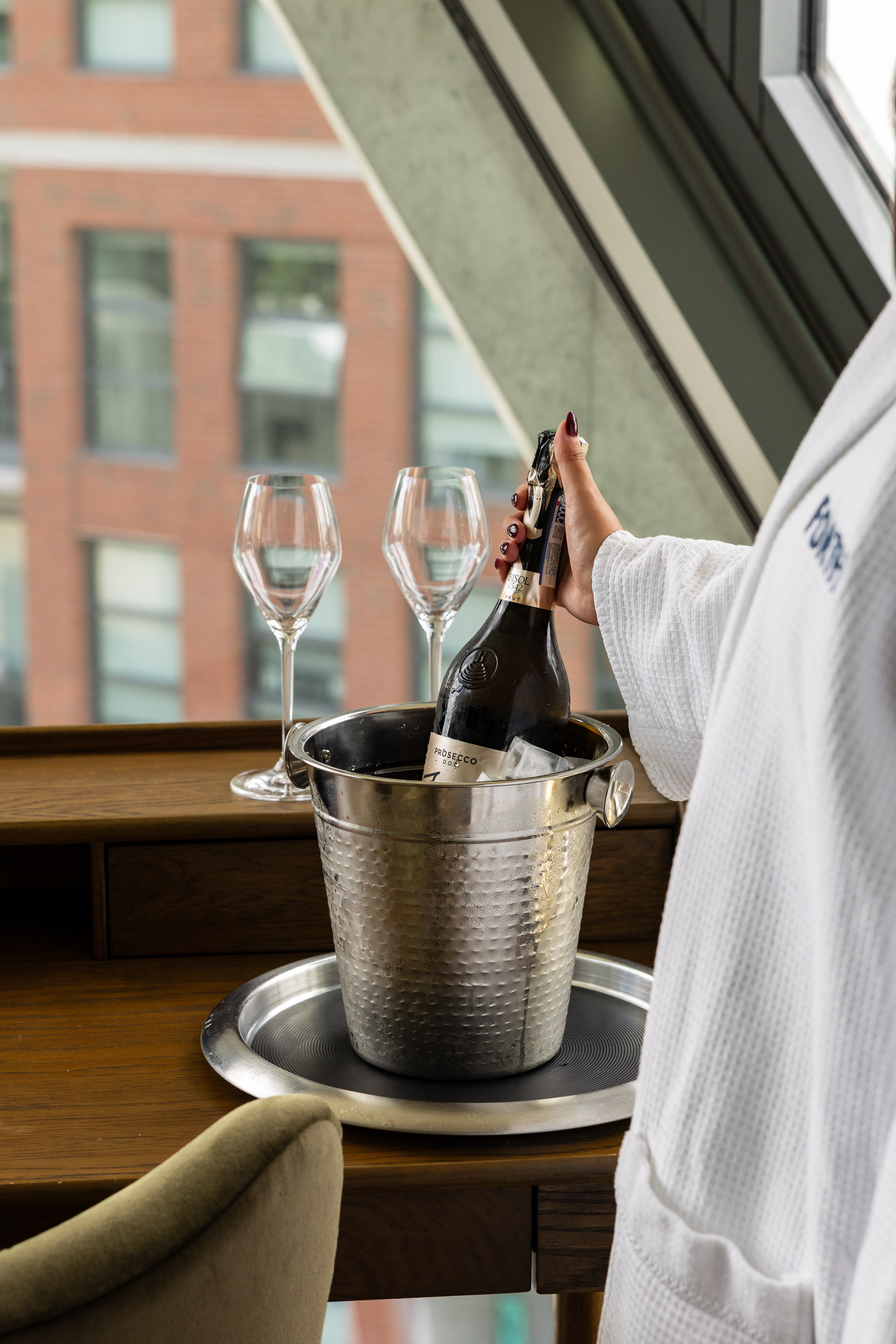 A woman pops a Prosecco bottle over ice in a metal bucket, with two wine glasses on a wooden table.