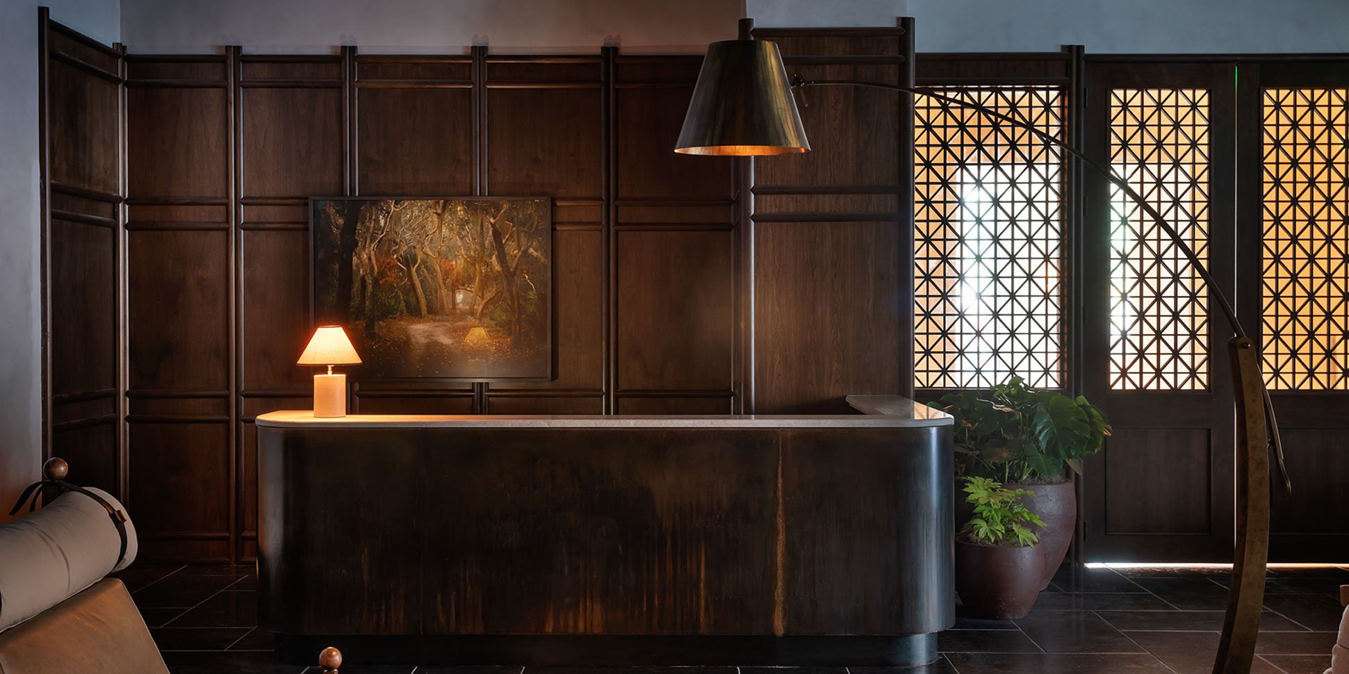 Dark wood reception room: desk with lamp, landscape painting, and patterned window with potted plants by it.